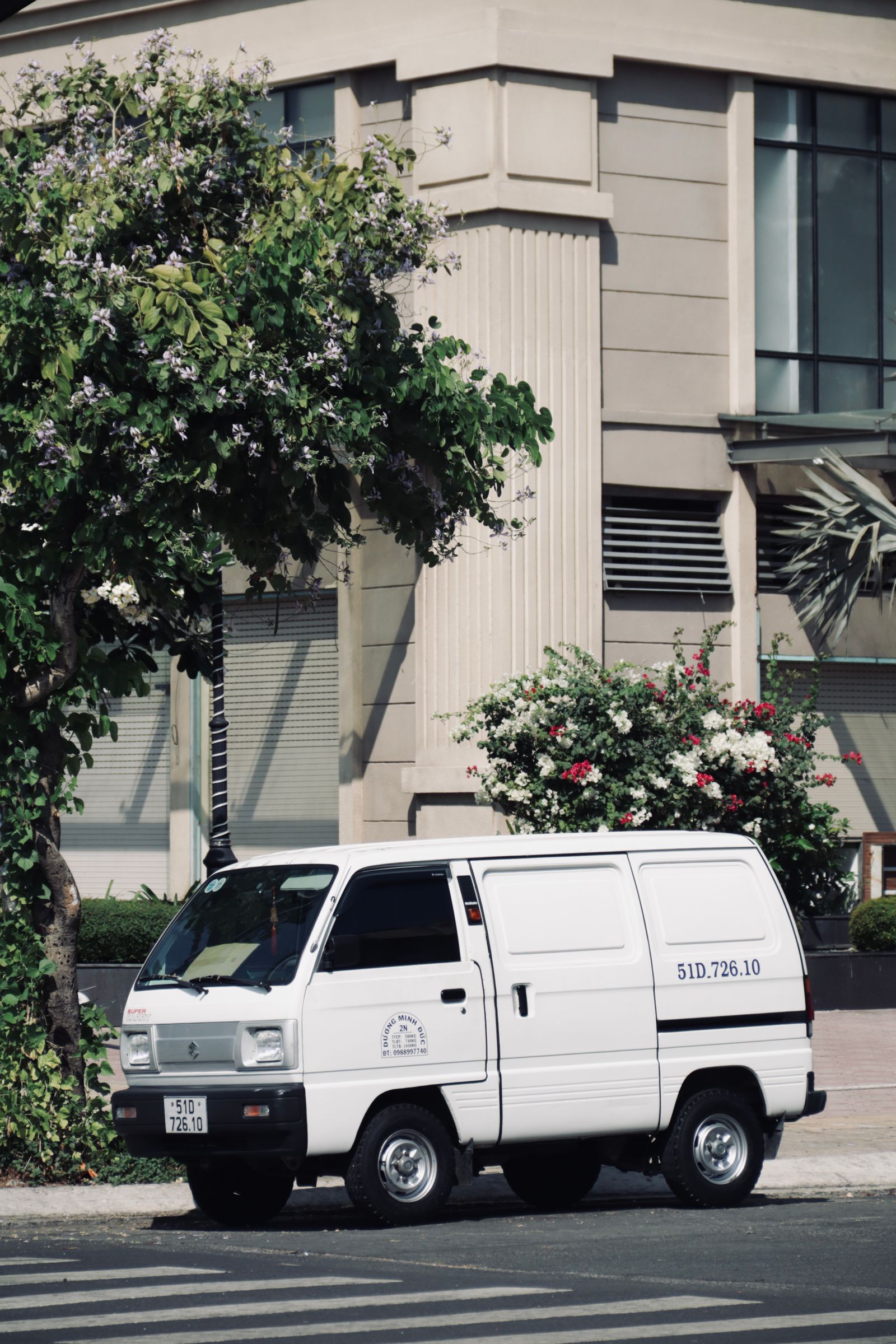 Small white compact cargo van parked beside a tan building with a flowering tree and shrubs, sunny, clean side/front 3/4 view, small license text on door but no overlay.