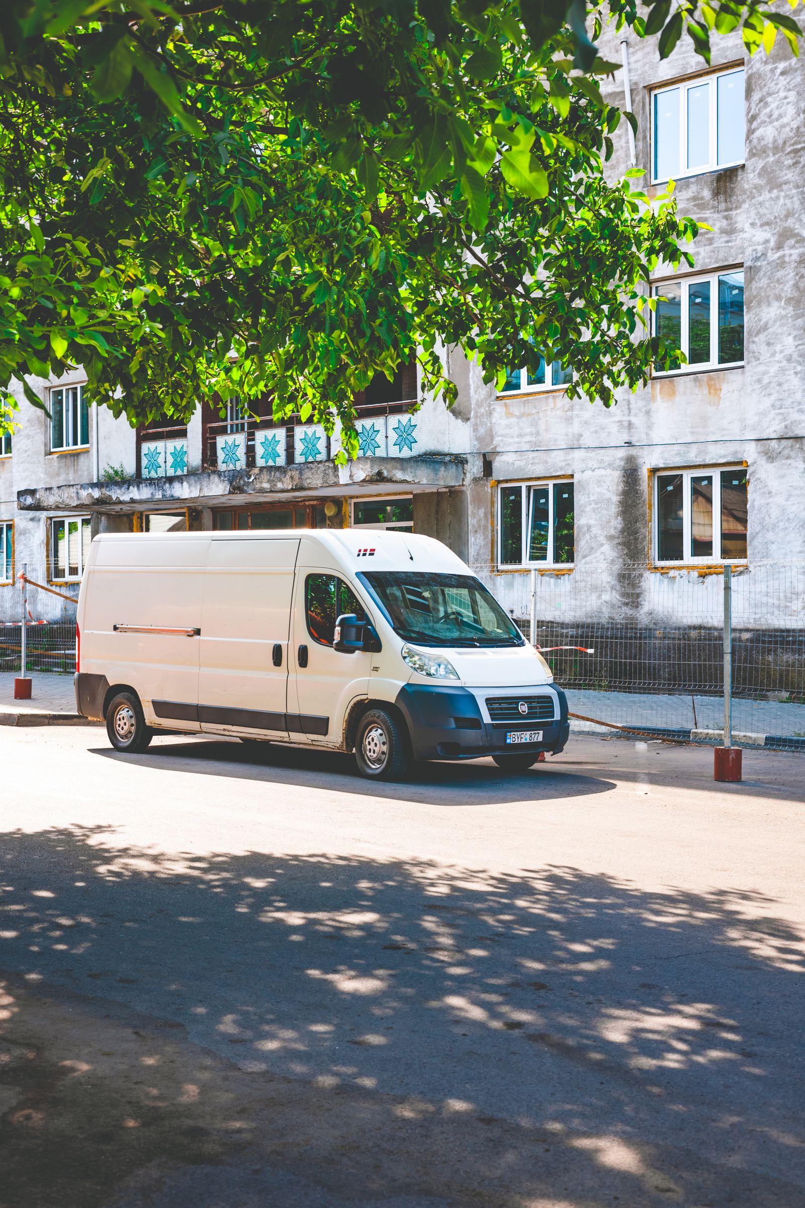 White Fiat Ducato high-roof cargo van parked on a sunny street under green tree shade, older building behind, clean 3/4 front view, no visible branding.