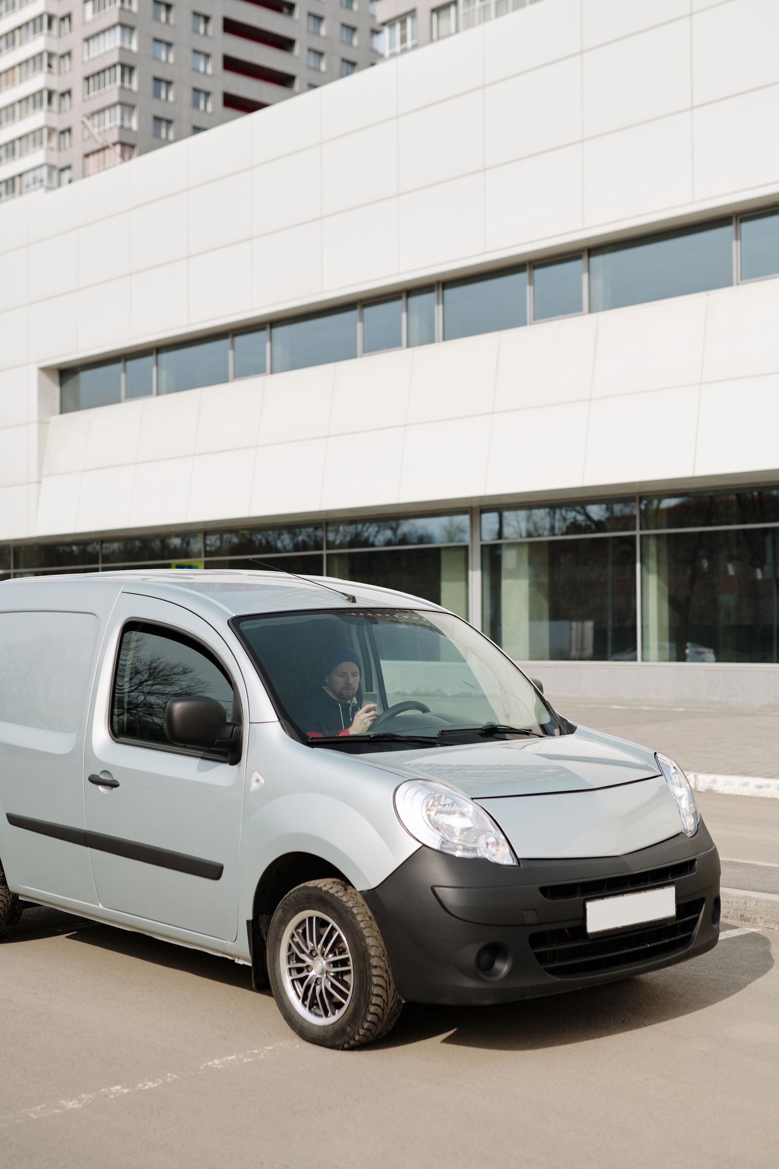 Light-silver compact cargo van parked in front of a multi-story commercial building, 3/4 front view, driver faintly visible inside, clean and modern.
