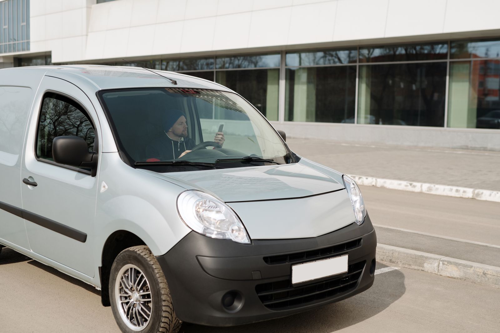 Light-silver compact cargo van, tight 3/4 front angle, glass-walled building behind, clean street, bright daylight. Blank front plate, no branding.