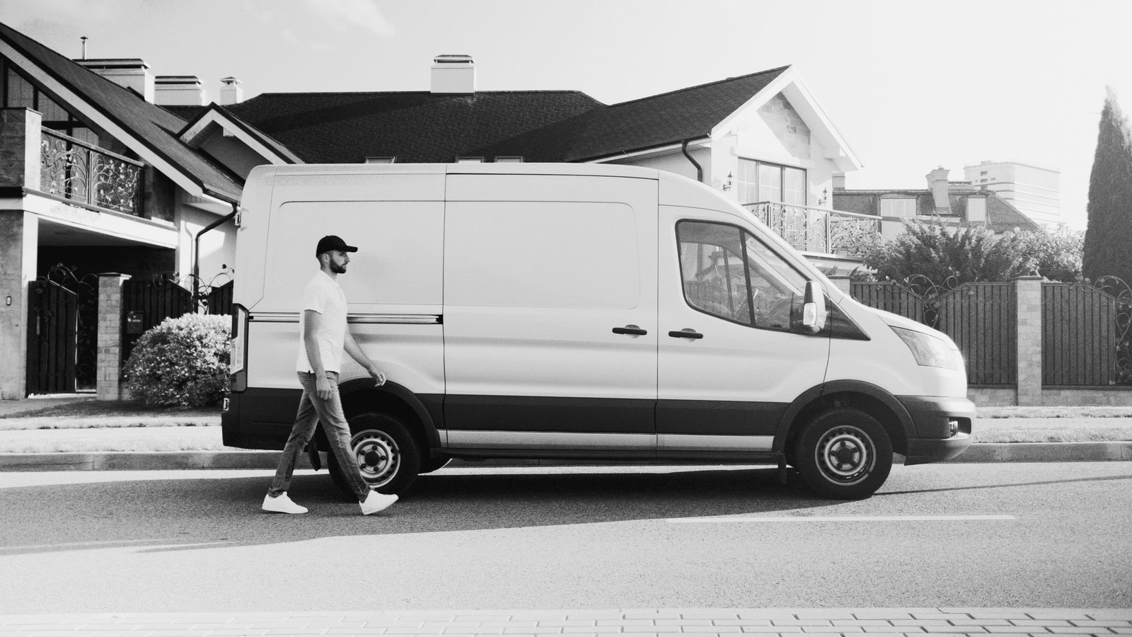 Black-and-white photo of a white Ford Transit cargo van parked on a residential street with houses behind; a man in cap and tee walks past it. Clean composition, monochrome.